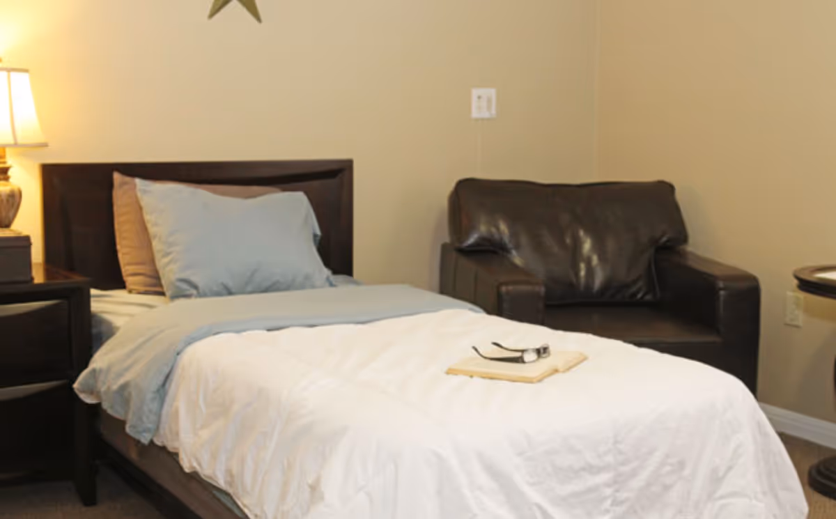 A bedroom with a single bed made with white and light blue bedding, two pillows, a dark wooden headboard, a nightstand with a lamp, a dark brown leather armchair, and a small round table. There is a book and a pair of glasses on the bed.