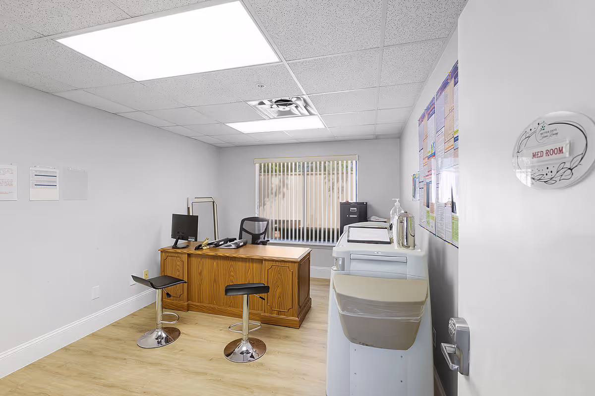 A small office room with a wooden desk, two black swivel stools, a computer monitor, and office supplies. There is a window with vertical blinds on the back wall, a filing cabinet, and a water dispenser with a trash bin attached. The door is partially open with a sign that reads 'MED ROOM'.