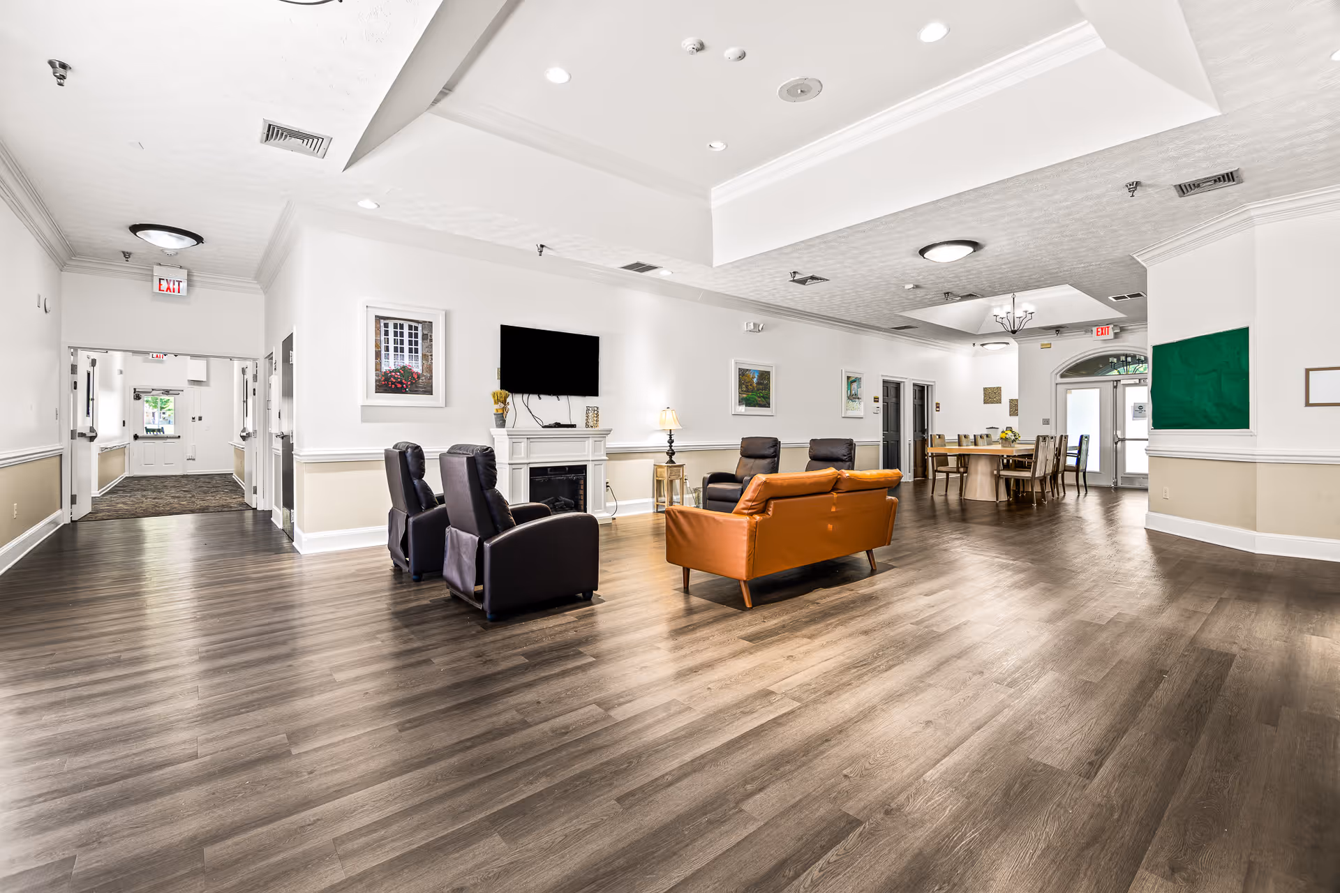 Spacious assisted living common area with sofas and recliners, a TV over a fireplace, and a dining table in the background under bright ceiling lights.