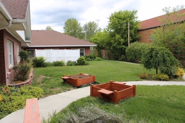 Outdoor garden area at Sarah Daft Home with green grass, two raised wooden garden beds, a concrete pathway, and surrounding bushes and trees. Buildings with brick walls and brown roofs are visible in the background under a partly cloudy sky.