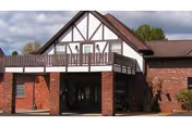 Exterior view of a senior living facility building with a brick facade and Tudor-style architectural elements, featuring a covered entrance and a balcony above it.