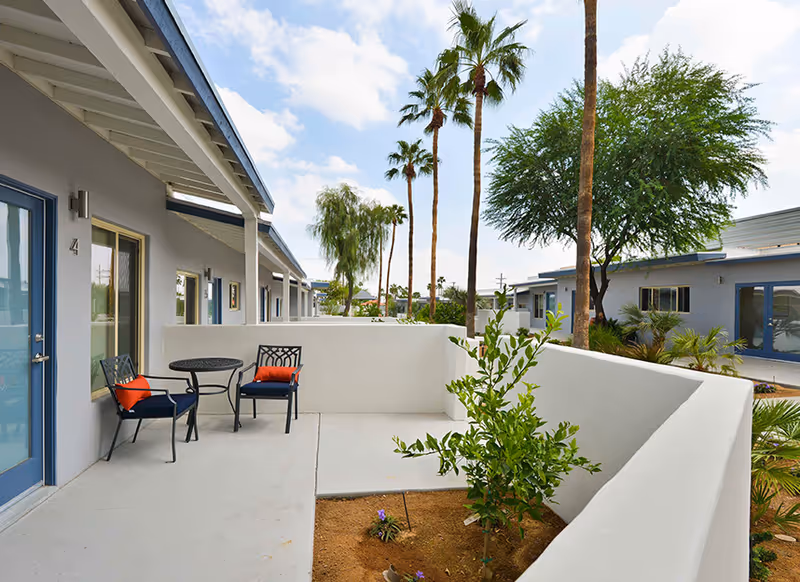 Outdoor patio area at Stonewall Gardens Assisted Living with two black metal chairs with orange cushions and a small round table. The patio is surrounded by white walls and overlooks a garden area with small plants and palm trees under a partly cloudy sky.