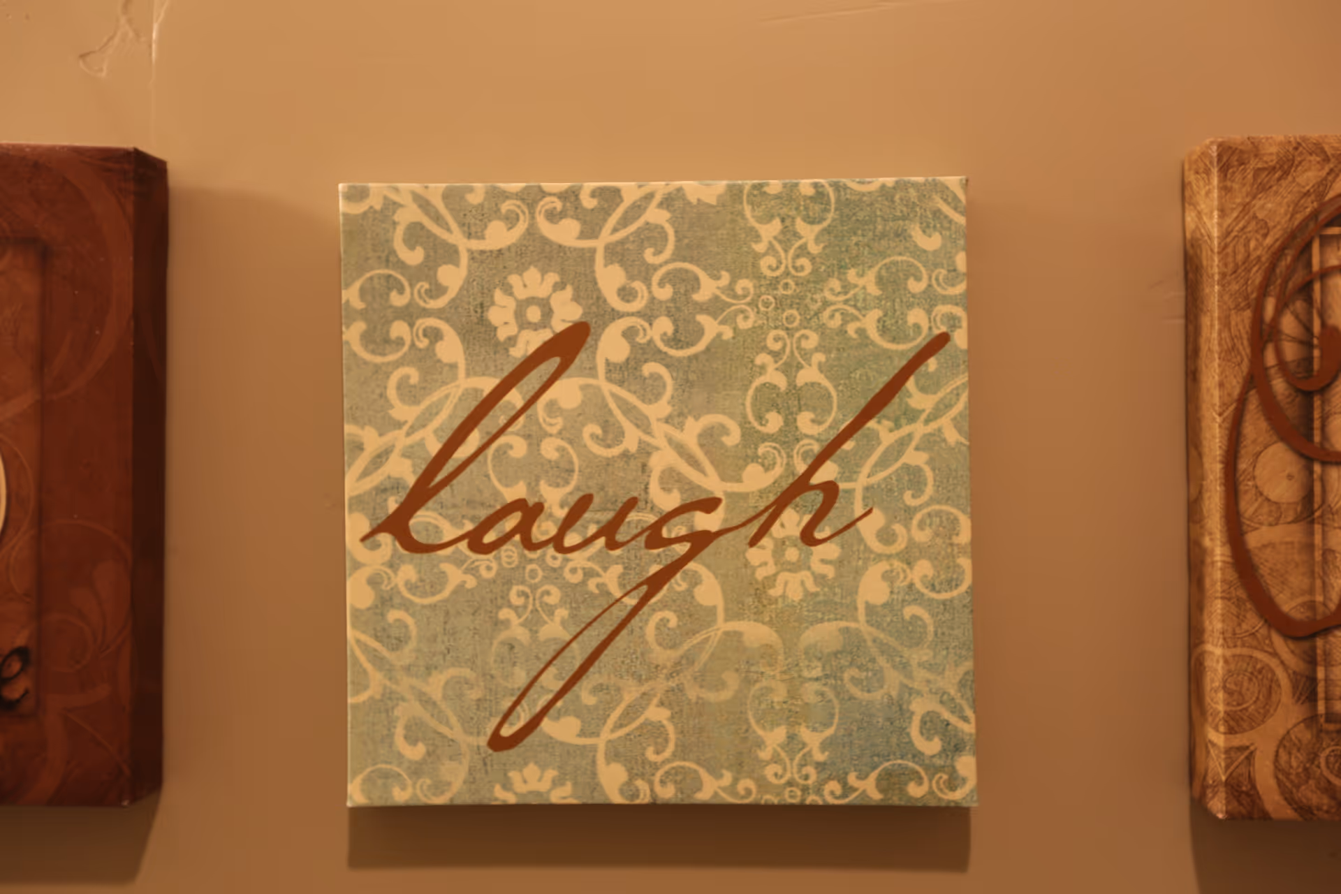 A decorative wall art piece with a blue and beige patterned background and the word 'laugh' written in cursive brown letters.