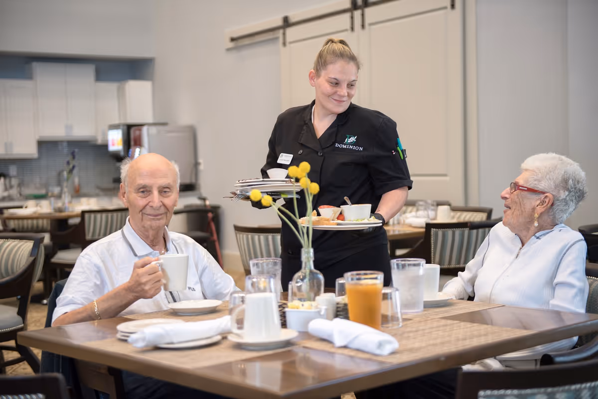 A senior man and woman sitting at a dining table in a senior living facility, with a staff member in a Dominion uniform serving food and drinks. The table has glasses of water, a glass of orange juice, a vase with yellow flowers, and neatly folded napkins. The background shows a kitchen area with white cabinets and appliances.