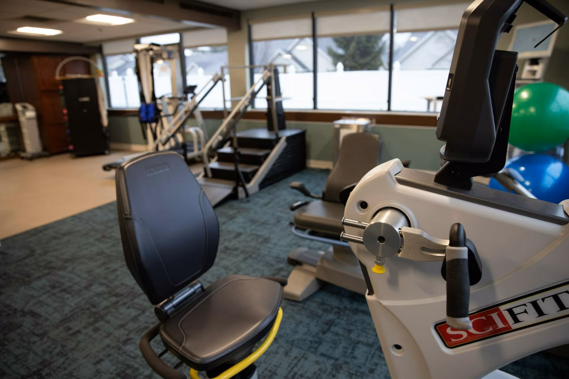 Interior view of a fitness room at Spring Mill Meadows featuring exercise equipment including a recumbent bike, resistance machines, and large exercise balls near windows with a view of houses outside.