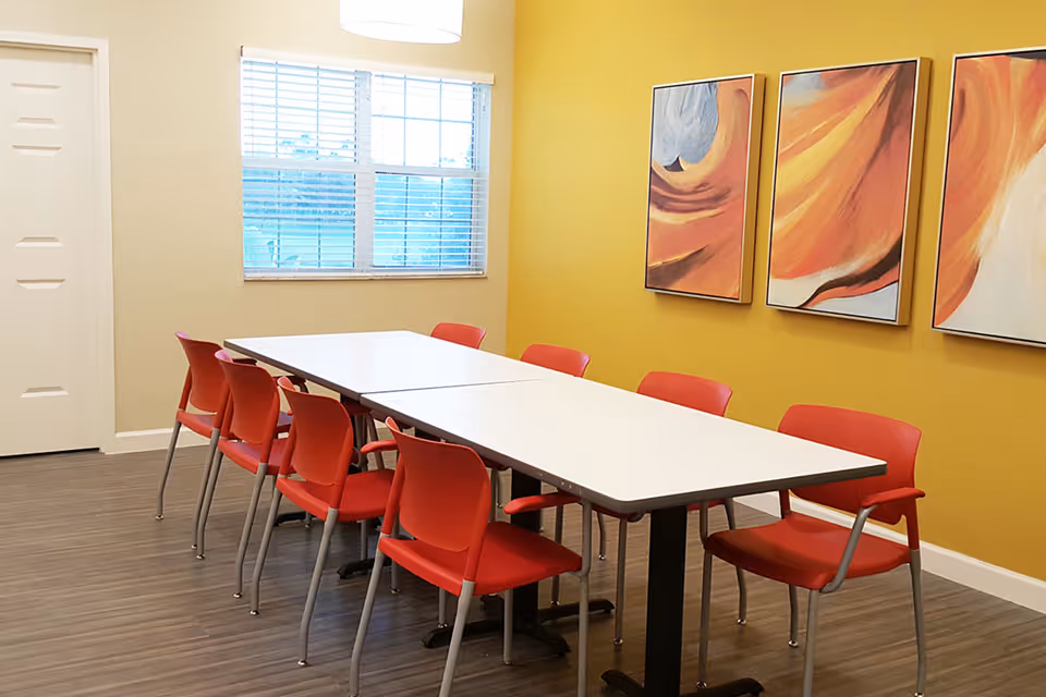 A bright dining/activity room with two white rectangular tables surrounded by red chairs, a yellow accent wall with abstract paintings, and a window.