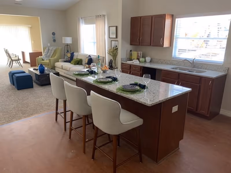 Interior view of a senior living facility showing a kitchen island with three white cushioned bar stools and place settings with plates, napkins, and wine glasses. Behind the island is a kitchen area with wooden cabinets, a double sink, and a window with blinds. In the background, there is a living room area with a white sofa, green armchair, coffee table, and additional seating near a window with curtains.