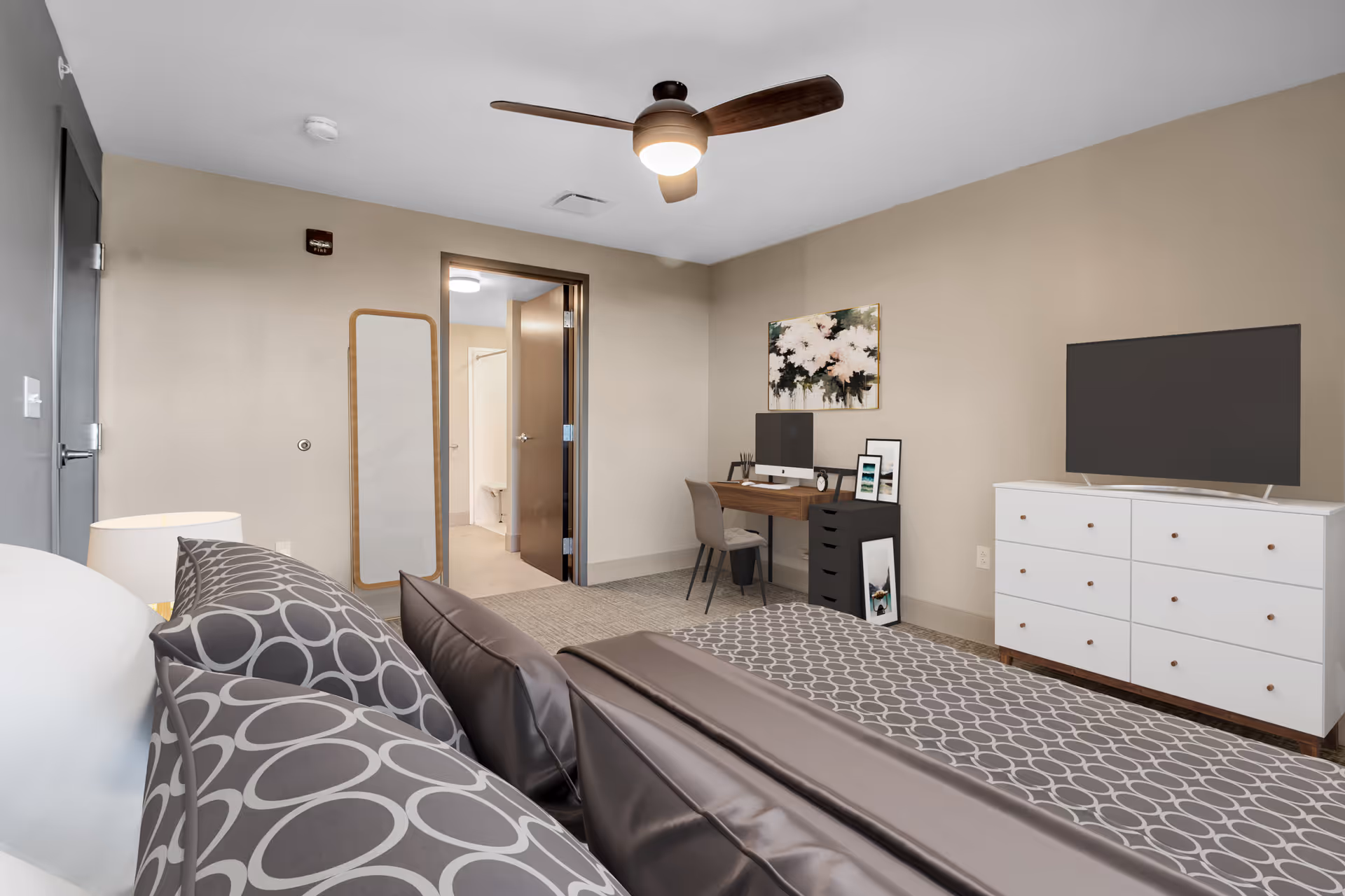 A modern bedroom with a bed featuring gray and white circular patterned bedding, a white dresser with a flat-screen TV on top, a wooden desk with a computer and chair, a full-length mirror on the wall, and a ceiling fan with a light. The room has beige walls and carpeted floor, with an open door leading to a bathroom.