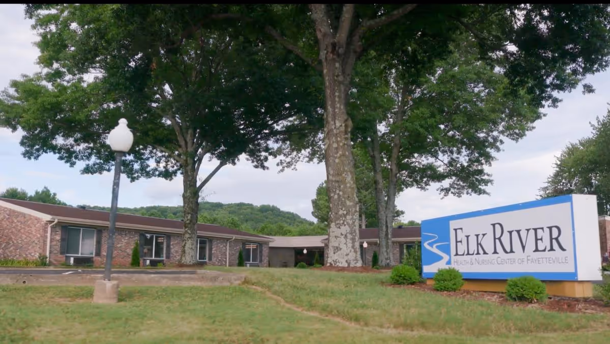 Exterior view of Elk River Health and Nursing Center of Fayetteville showing a single-story brick building with large trees and a grassy lawn. A white and blue sign with the facility name is visible in the foreground.