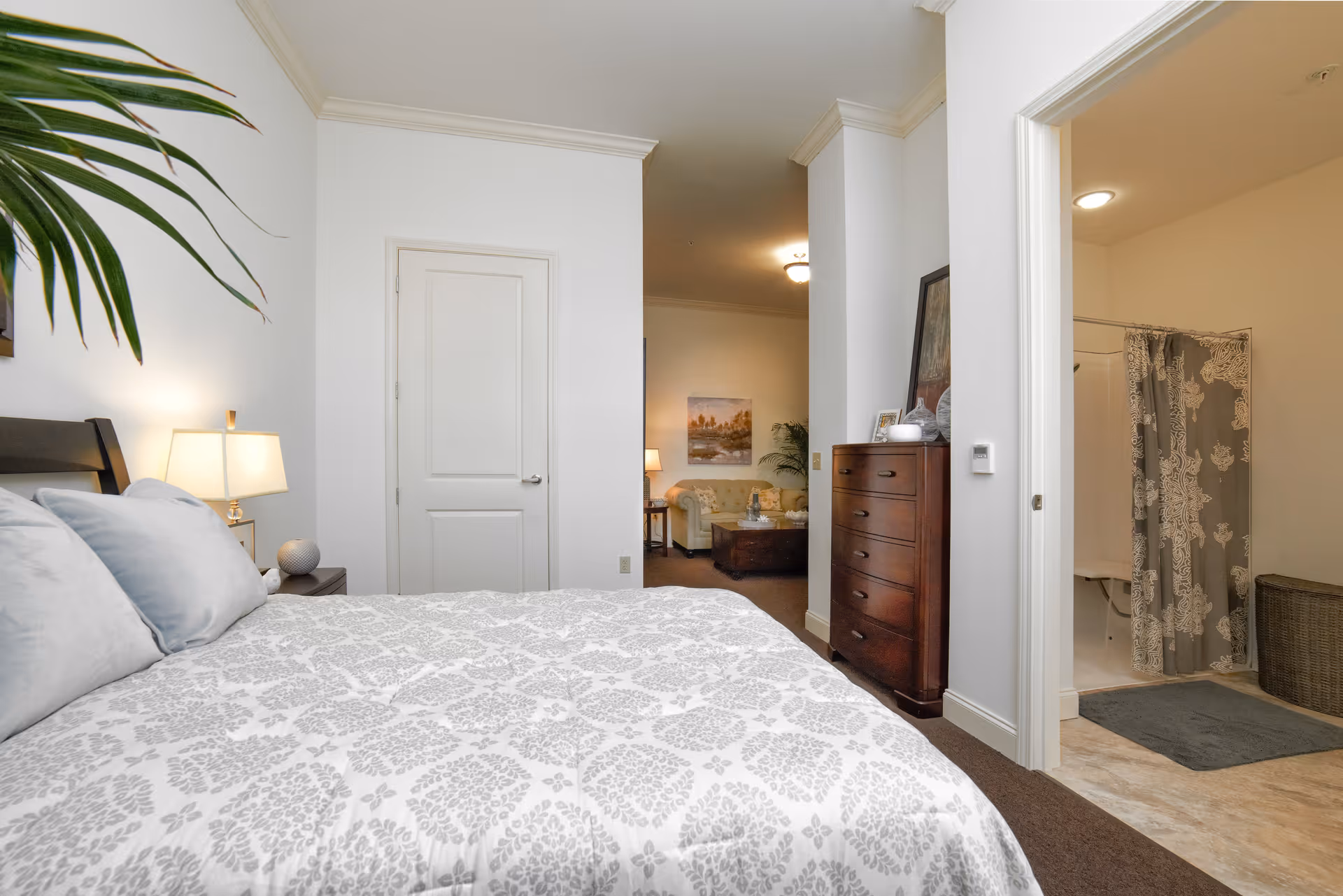 A bedroom with a bed covered in a white and gray patterned comforter and pillows. A bedside table with a lamp and decorative items is next to the bed. A wooden dresser stands against the wall near an open doorway leading to a bathroom with a shower and a gray patterned shower curtain. In the background, a living room area with a sofa, coffee table, lamp, and wall art is visible.