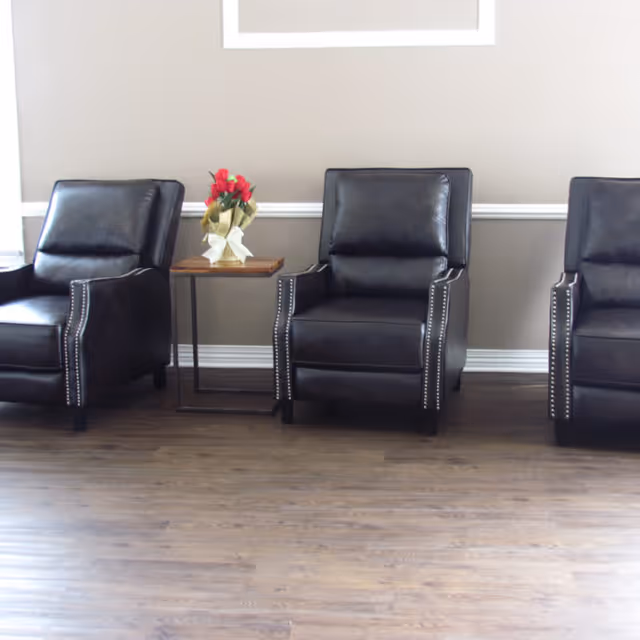 Three black leather armchairs arranged side by side against a beige wall with white trim. A small wooden side table with a metal frame sits between the first and second chairs, holding a bouquet of red flowers wrapped in gold and white paper. The floor is a medium-tone wood.