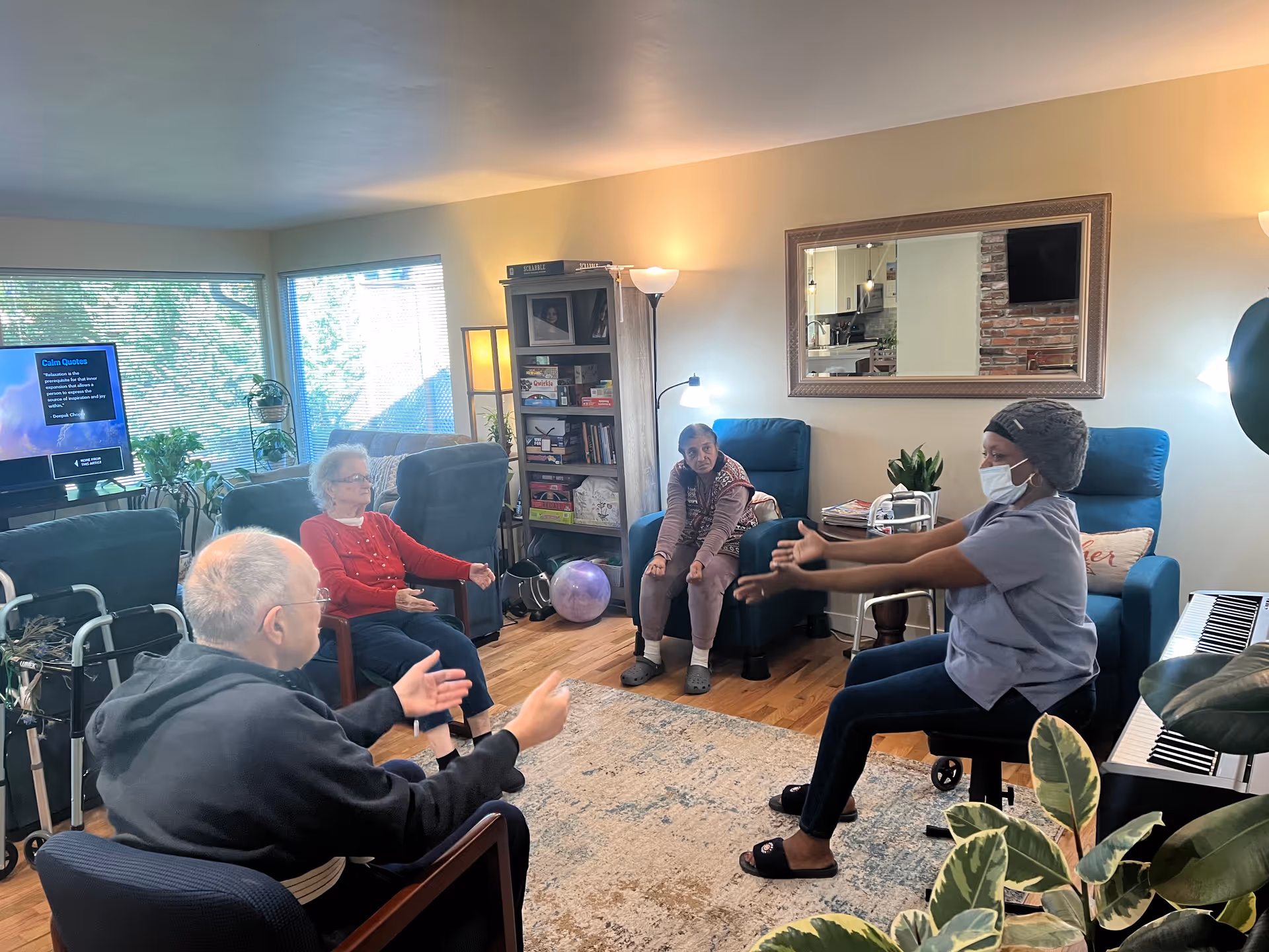 A group of elderly individuals and a caregiver wearing a mask are seated in a cozy living room with wooden floors, blue armchairs, a bookshelf, and a large mirror on the wall. The caregiver is leading an exercise or stretching activity while the elderly participants follow along. There is a television in the background displaying calm quotes, and the room is well-lit with natural light coming through the windows.