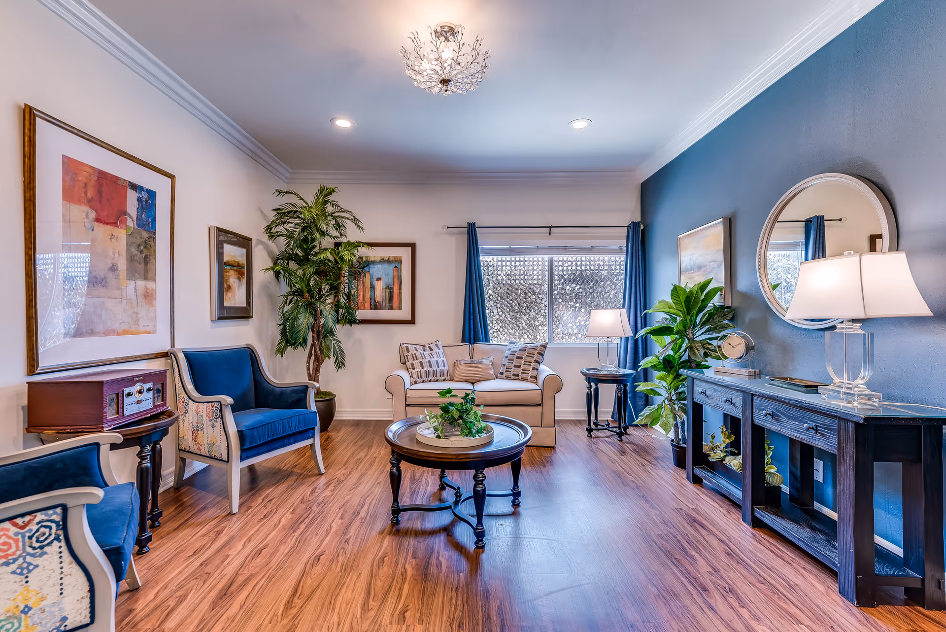 A bright and cozy senior living room with wooden flooring, a beige loveseat with patterned pillows, two blue upholstered armchairs, a round wooden coffee table with a plant centerpiece, a side table with a vintage radio, a console table with a lamp and clock, a round mirror on a blue accent wall, framed artwork, and large window with blue curtains.