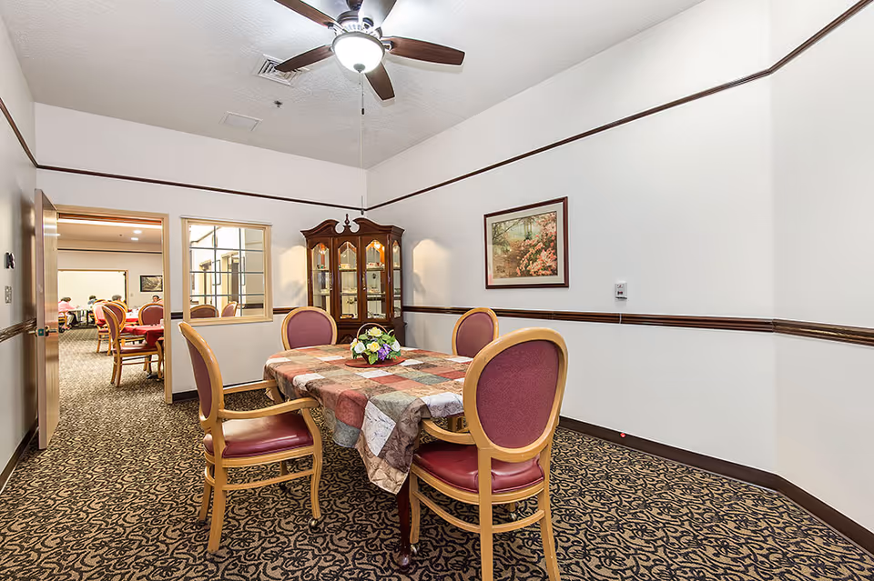 A small dining area with a table covered by a patchwork tablecloth and four wooden chairs with red cushions. A ceiling fan with a light is above the table. A wooden china cabinet with glass doors is against the wall, and a framed picture of a garden scene hangs on the wall. Through an open doorway, another room with more tables and chairs is visible.