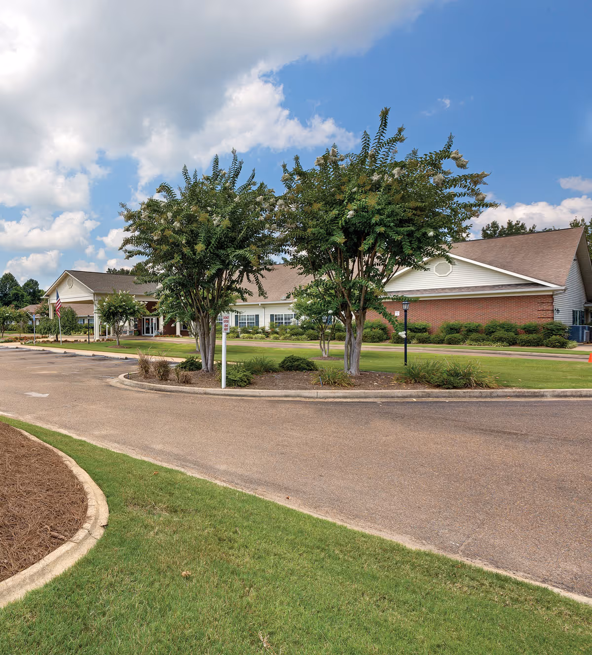 Exterior view of The Pinnacle of Oxford senior living facility showing a paved driveway, green lawn, trees, and a building with a brick and white facade under a partly cloudy blue sky.