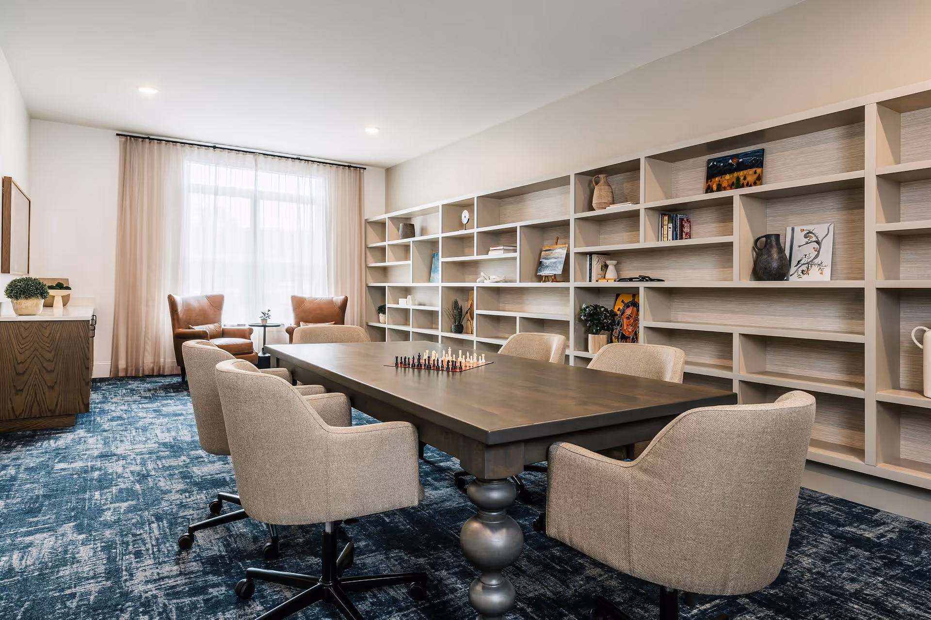 A bright communal activity room with a large wooden table surrounded by upholstered chairs, built-in bookshelves along one wall, and two armchairs by a window.