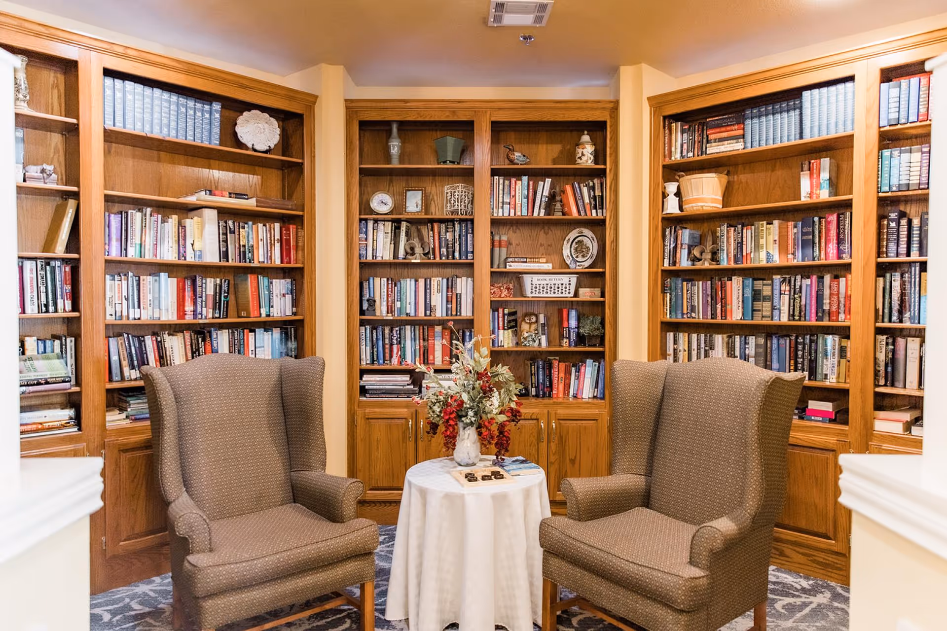 Two upholstered armchairs face a small round table with a floral arrangement, set in front of built-in wooden bookshelves filled with books and decor.