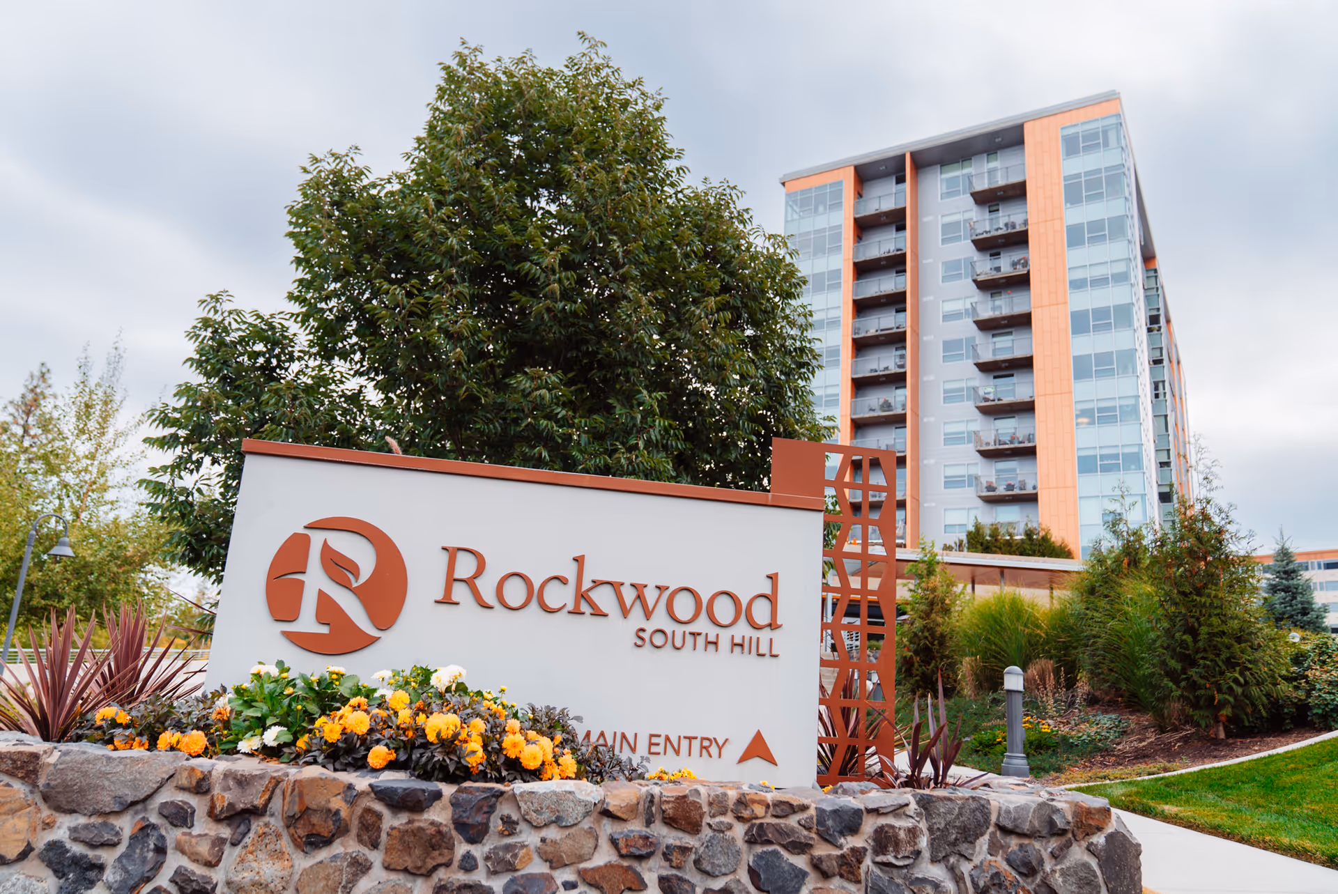Outdoor view of the Rockwood South Hill senior living facility sign with a landscaped garden featuring yellow and white flowers in front. Behind the sign, there is a multi-story building with balconies and large windows under a cloudy sky.
