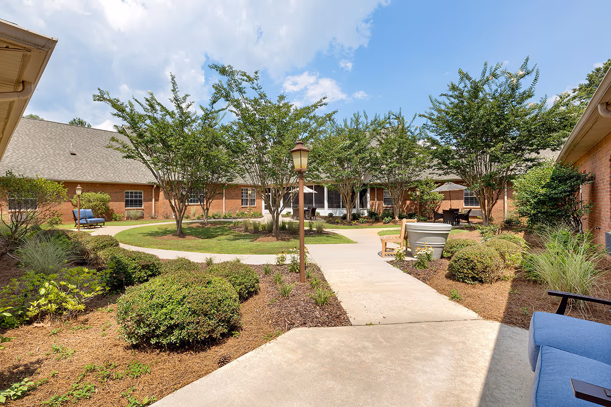 Outdoor courtyard area with a concrete pathway surrounded by green bushes and trees. There are benches and patio tables with umbrellas placed around the courtyard. The buildings surrounding the courtyard have brick walls and multiple windows under a partly cloudy sky.