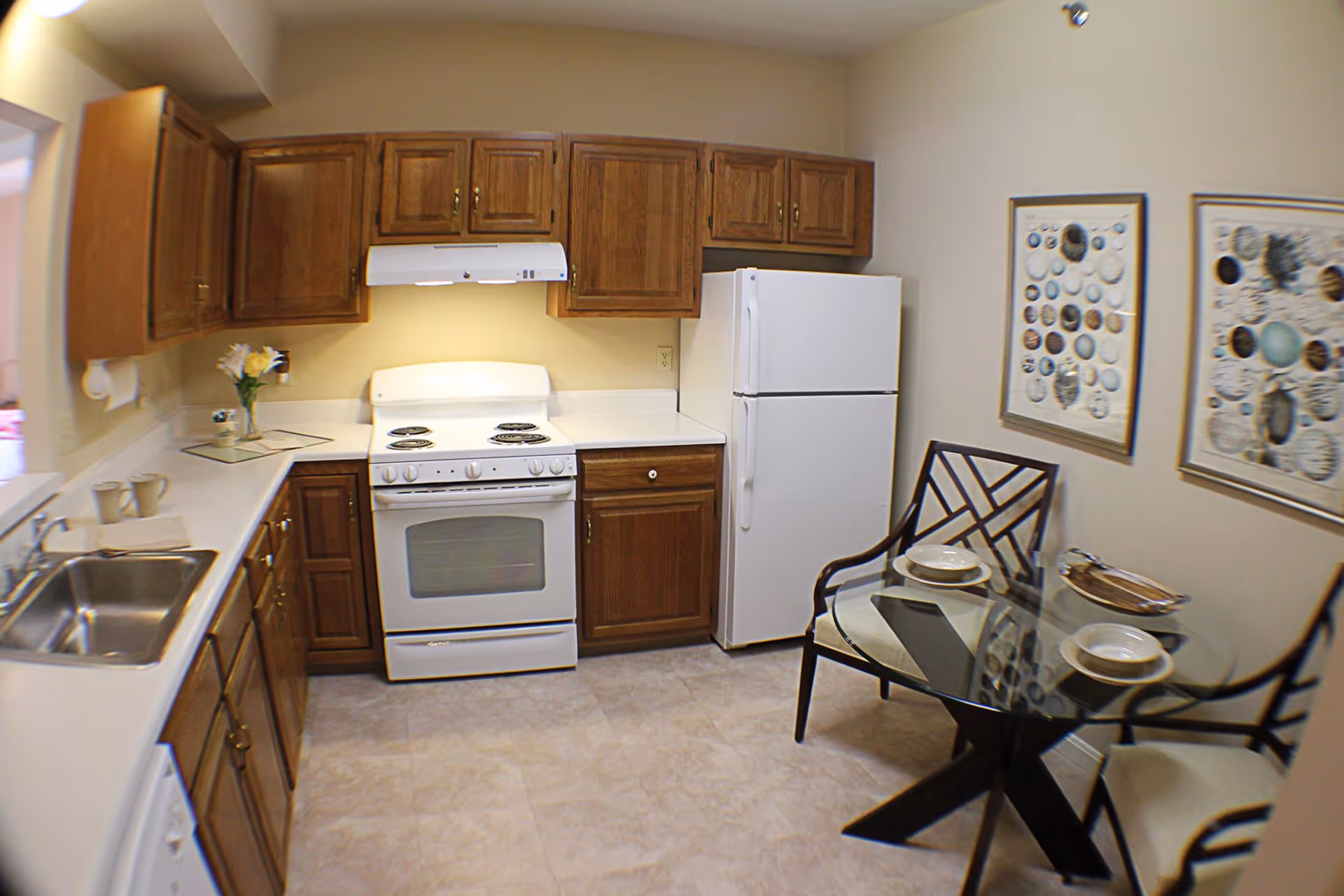 A kitchen with wooden cabinets, a white stove with four burners, a white refrigerator, and a stainless steel sink. There is a small glass dining table with two chairs, each set with a bowl and plate. Two framed pictures with shell designs hang on the wall above the dining area.