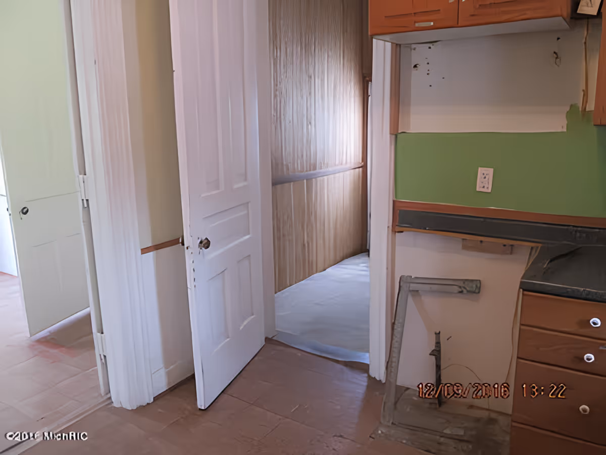 Interior view of a partially renovated room with an open white door leading to a narrow hallway with wood paneling. The room has a green wall with an electrical outlet and wooden cabinets above a countertop area that appears to be under construction or repair. The floor is covered with a protective sheet in the hallway and the timestamp 12/19/2016 13:22 is visible in the bottom right corner.