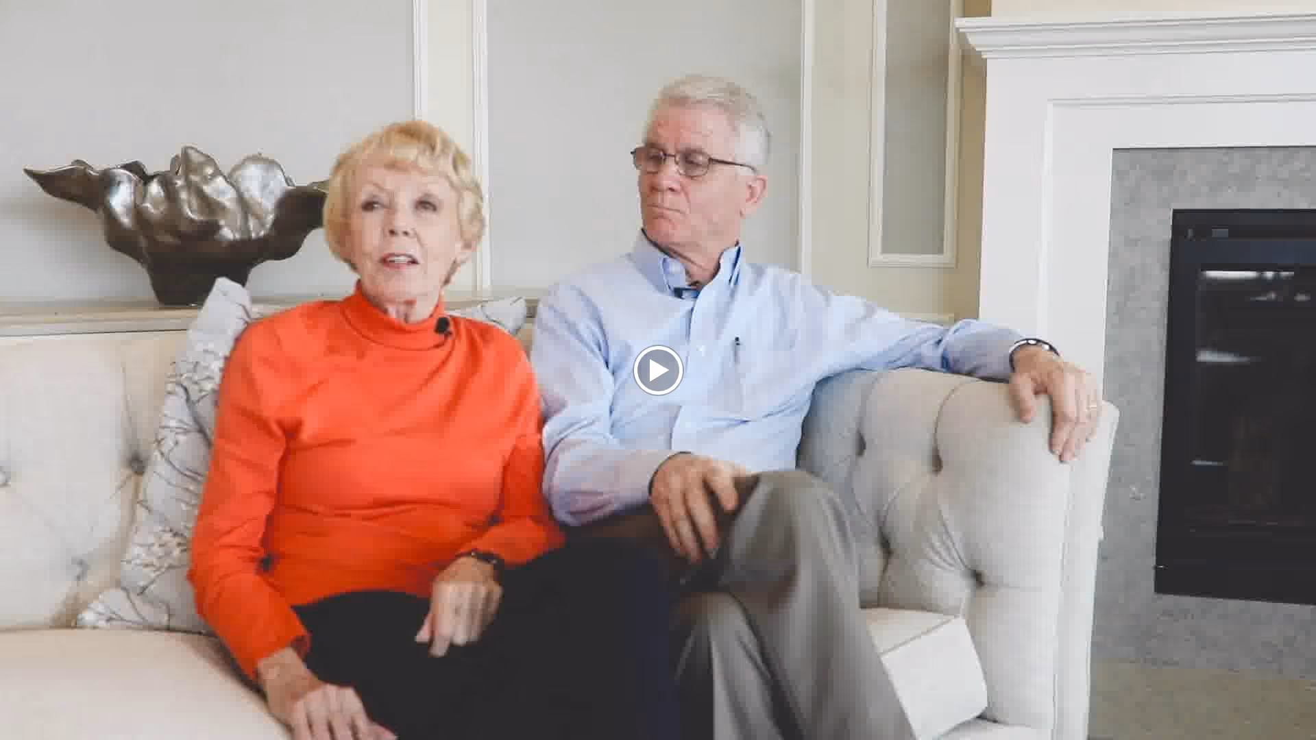 An elderly couple sitting on a light-colored tufted sofa in a living room. The woman is wearing an orange long-sleeve top and the man is wearing a light blue button-up shirt with glasses. Behind them is a decorative sculpture and a fireplace is visible to the right.