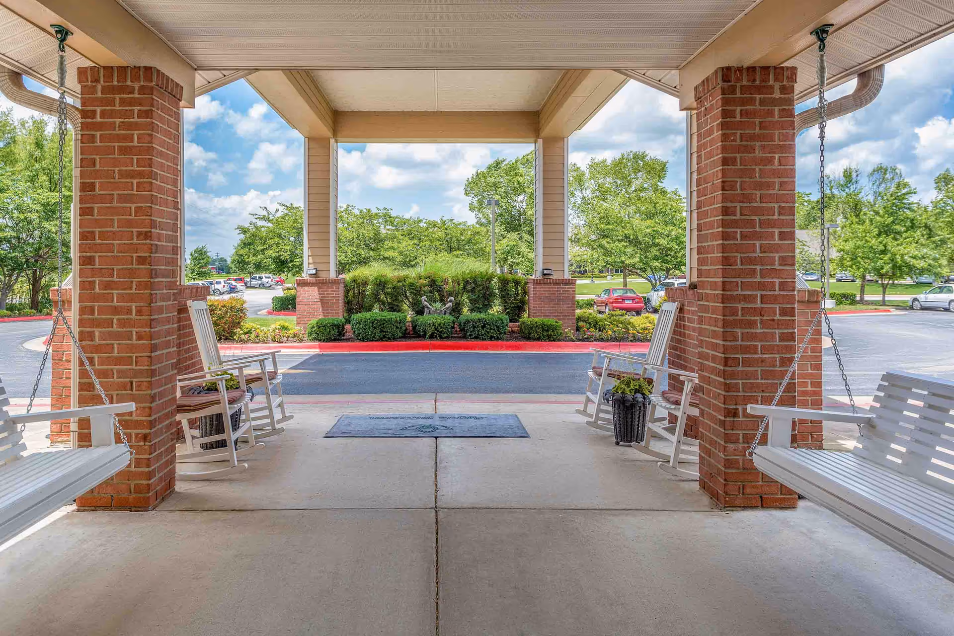 Covered outdoor entrance area with two brick columns, white wooden swings hanging from the ceiling on both sides, and white rocking chairs with cushions. Beyond the entrance is a parking lot with cars and green trees under a partly cloudy sky.