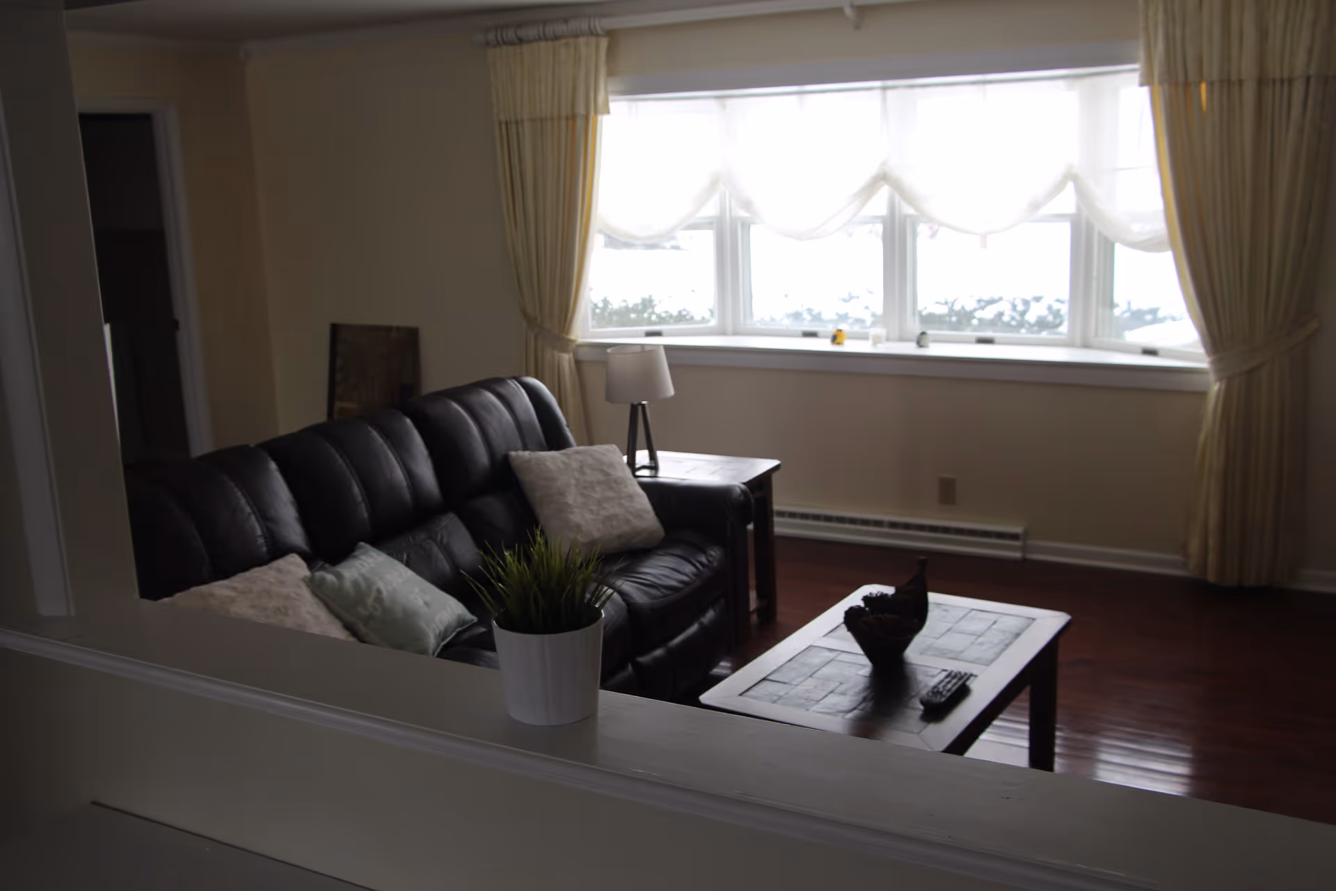 A cozy living room with a black leather couch adorned with pillows, a wooden coffee table with decorative items and a remote control, a side table with a lamp, and a large window with sheer white curtains and beige drapes letting in natural light.