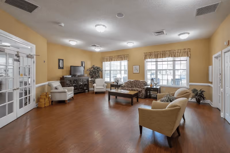 A spacious living room with yellow walls and wooden flooring, furnished with a floral-patterned sofa, four beige armchairs, a dark wooden coffee table, and a TV on a cabinet. Large windows with valances allow natural light to fill the room, and there are decorative plants and framed artwork on the walls.