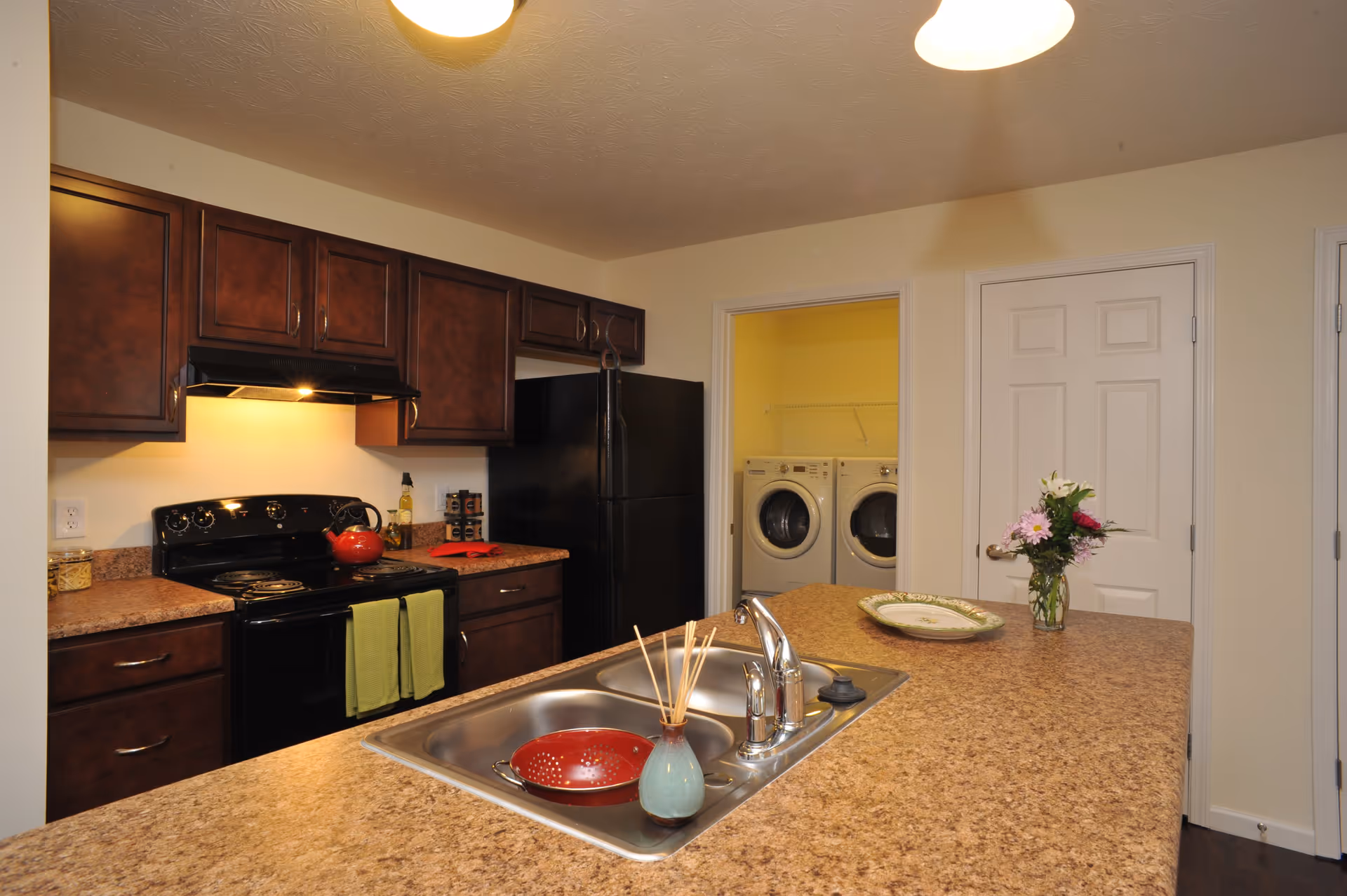 Interior view of a kitchen with dark wooden cabinets, a black stove with a red kettle, a black refrigerator, and a countertop with a sink. In the background, a laundry area with a washer and dryer is visible through an open door. There is a vase with flowers and a decorative plate on the countertop.