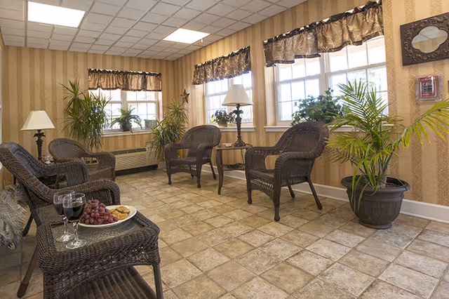 Sunlit sitting room with wicker chairs, side tables with lamps and snacks, and potted plants by windows.