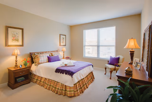 A cozy bedroom in a senior living community featuring a bed with plaid bed skirt, white bedding, and purple pillows and throw. There are two bedside tables with lamps and framed artwork above the bed. A window with blinds lets in natural light. A wooden dresser with a lamp, framed photos, and decorative items is on the right side, along with an upholstered chair. A green plant is visible in the foreground.