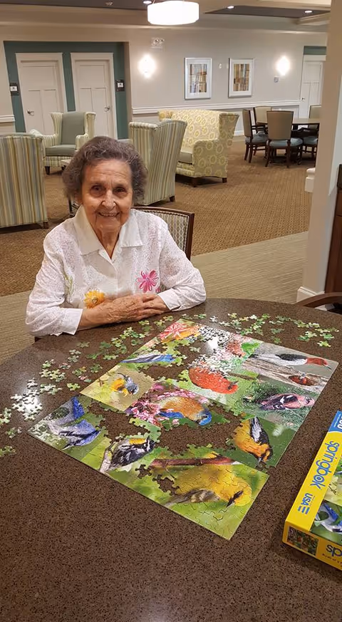 An elderly woman with short gray hair wearing a white blouse with floral embroidery is sitting at a table working on a colorful bird-themed jigsaw puzzle in a well-lit common area with upholstered chairs and tables in the background.
