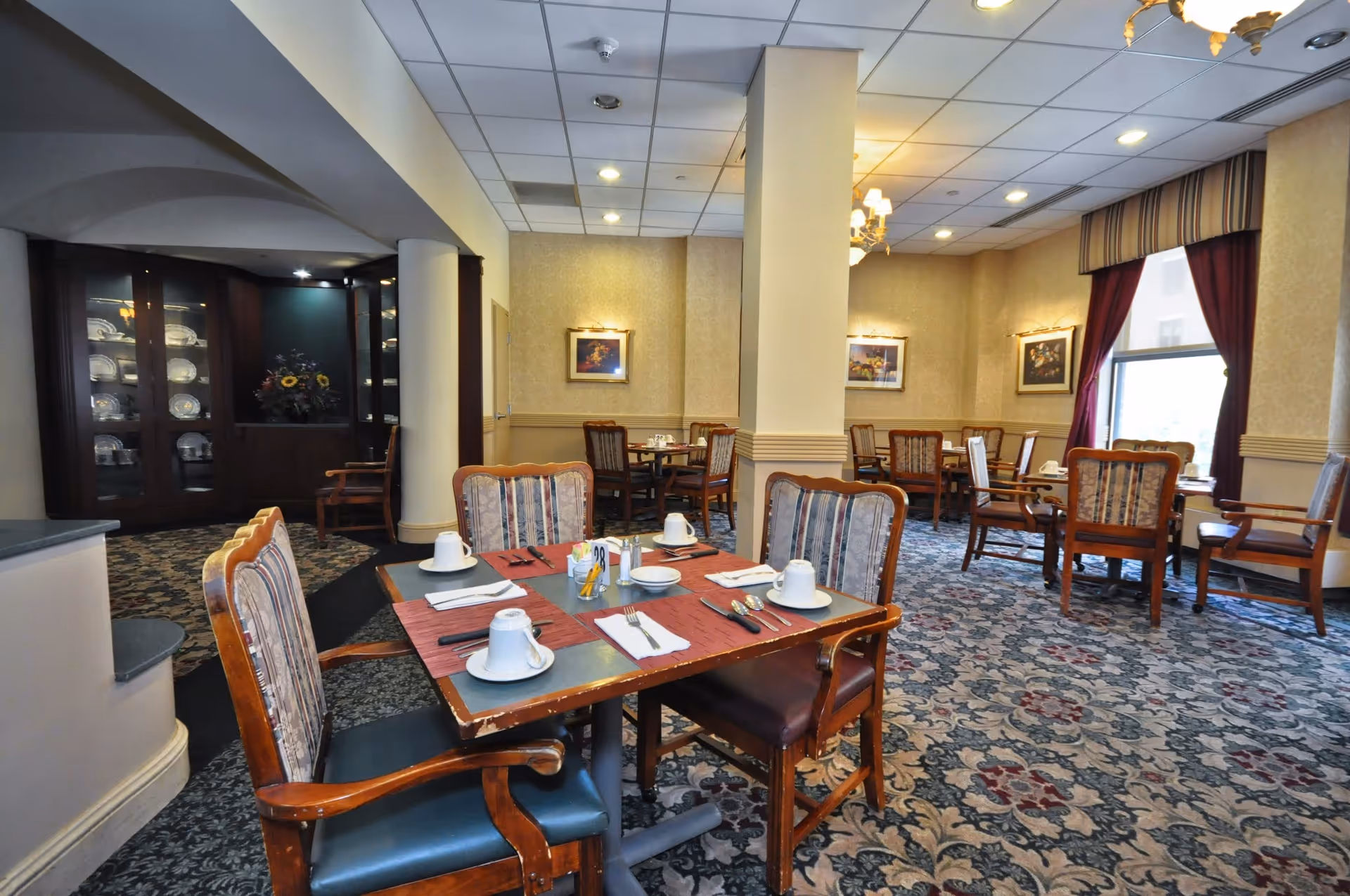 A dining room in St. Joseph Manor with several wooden tables and chairs arranged neatly. Each table is set with white cups, plates, utensils, and napkins. The room features patterned carpet, beige walls with framed artwork, and large windows with red curtains allowing natural light to enter. A cabinet displaying plates and a floral arrangement is visible in the background.