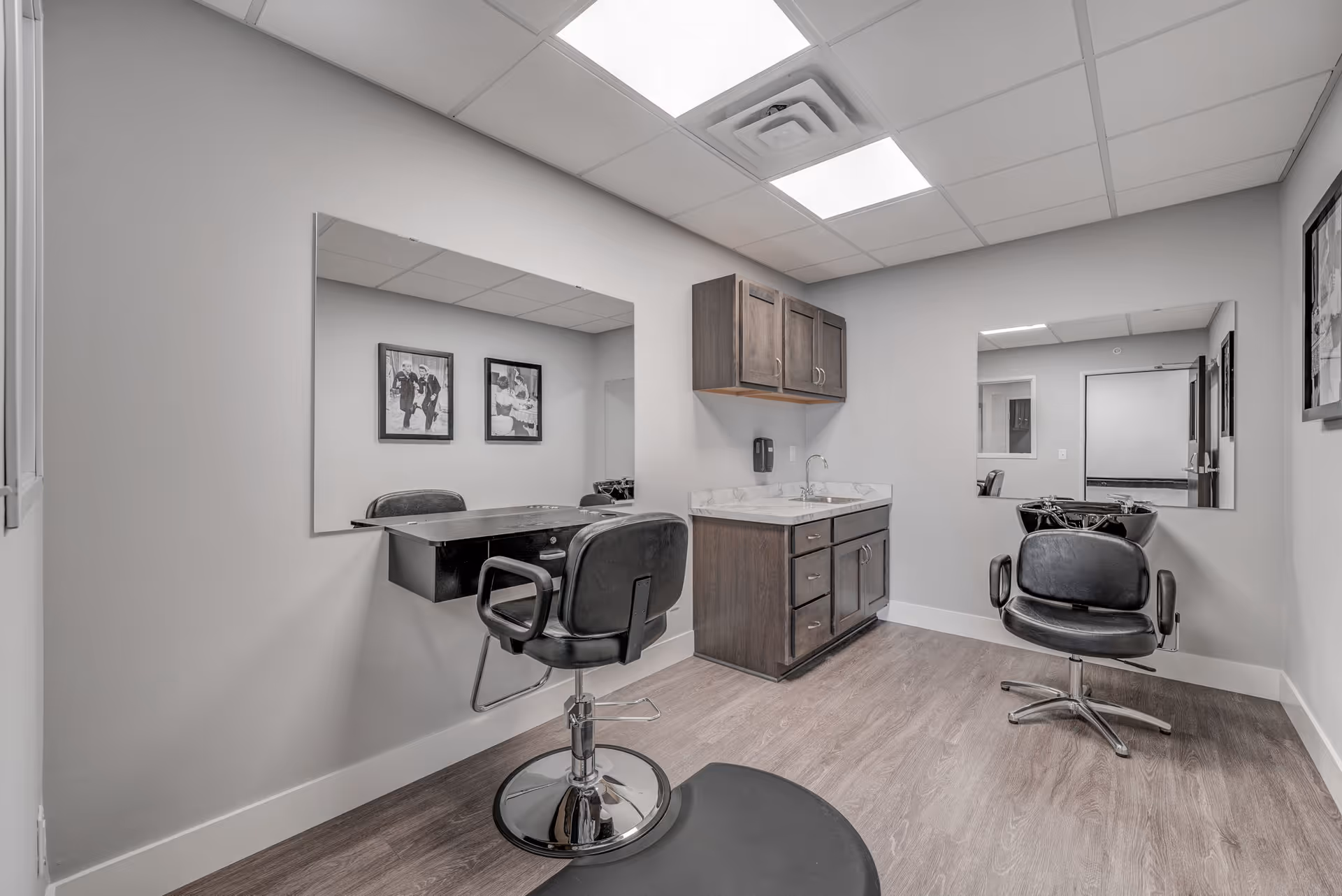 Interior view of a small salon room in a senior living facility with two black salon chairs, large wall mirrors, a wooden cabinet with a sink, and framed black and white photos on the walls.