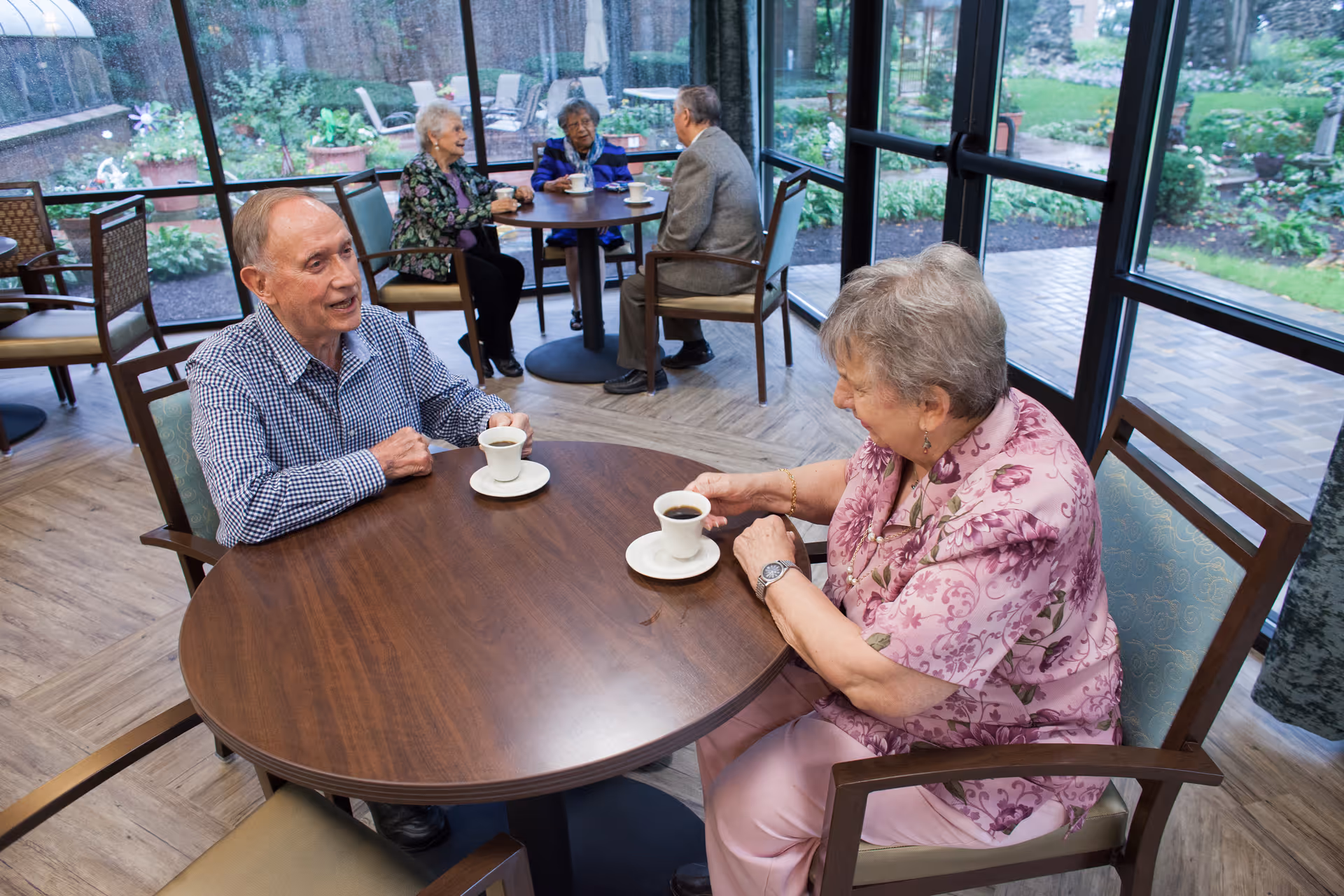 Two elderly people sitting at a round wooden table drinking coffee and talking inside a sunroom with large windows overlooking a garden. In the background, another elderly couple is seated at a similar table also enjoying coffee and conversation.