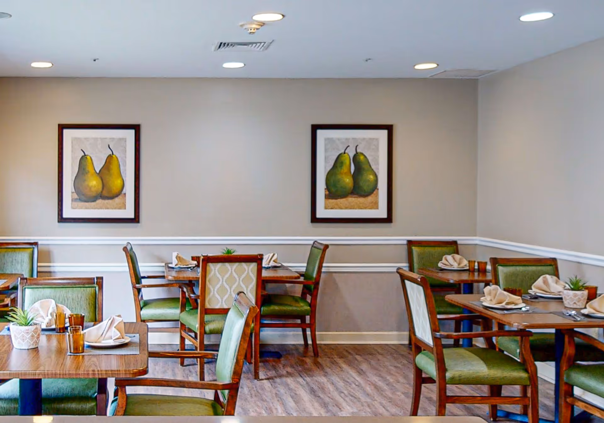 A dining room with wooden tables and green cushioned chairs. Each table is set with folded napkins, plates, and glasses. The walls are light-colored with white trim, and two framed paintings of pears hang on the wall. The floor is wooden, and ceiling lights illuminate the room.