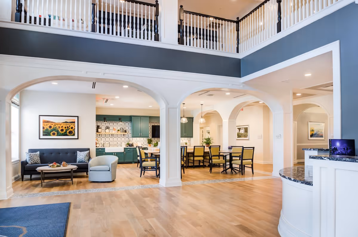 Bright and spacious interior of a senior living facility featuring a sitting area with a dark blue sofa, light blue armchair, and a coffee table. Adjacent to the sitting area is a dining space with multiple tables and chairs, green cabinetry, and a patterned backsplash in the kitchen area. The room has high ceilings with a balcony railing above and light wood flooring throughout.