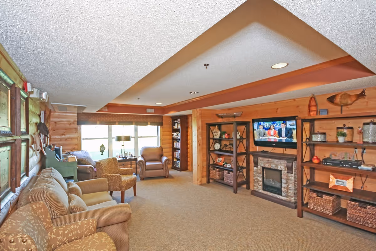 Cozy wood-paneled common living room with sofas and armchairs, shelving, a wall-mounted TV above a fireplace, and large windows.