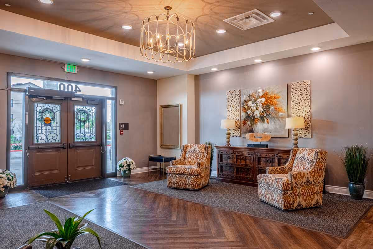 A well-lit lobby area with double glass doors featuring decorative ironwork. The floor has a combination of wood and carpet. Two patterned armchairs are placed on a carpet in front of a wooden sideboard with two table lamps and a floral painting above it. There are potted plants and a large mirror on the walls, with a decorative chandelier hanging from the ceiling.