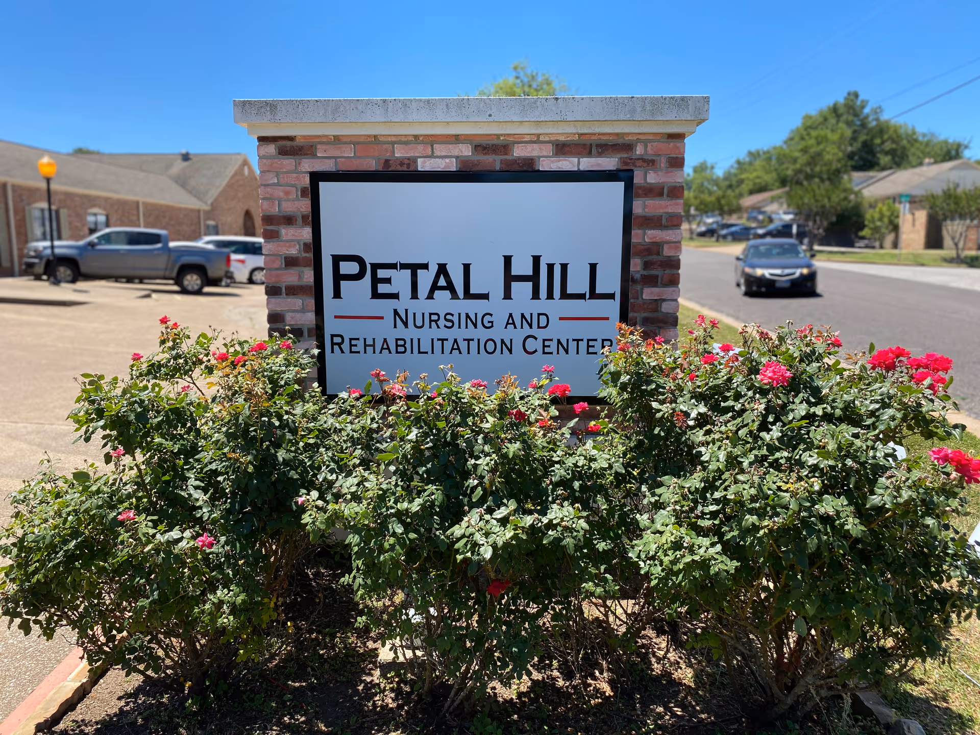Brick entrance sign reading "Petal Hill Nursing and Rehabilitation Center" surrounded by flowering bushes with a street and parked cars in the background.