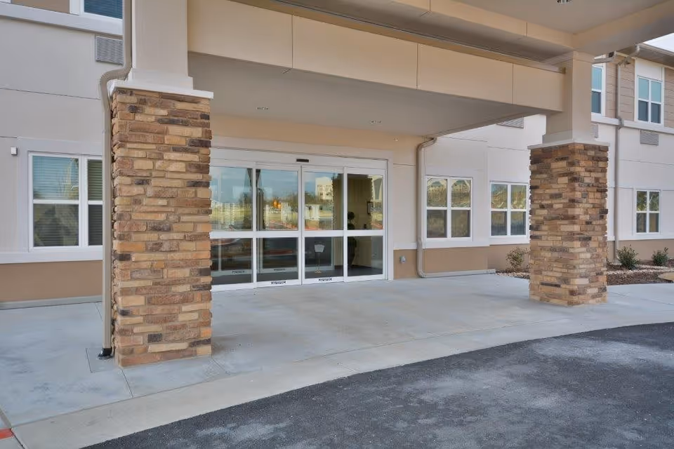 Entrance to a building with automatic sliding glass doors, supported by two pillars covered in decorative stone. The building exterior is beige with multiple windows, and there is a paved driveway in front.