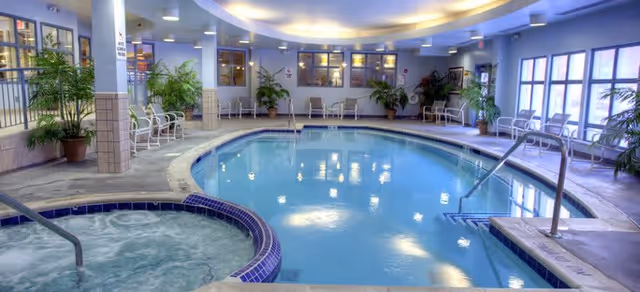 Indoor swimming pool area with a hot tub in the foreground. The pool is surrounded by tiled flooring and several potted plants. There are white chairs along the walls and large windows letting in natural light. The ceiling has recessed lighting and a curved design.