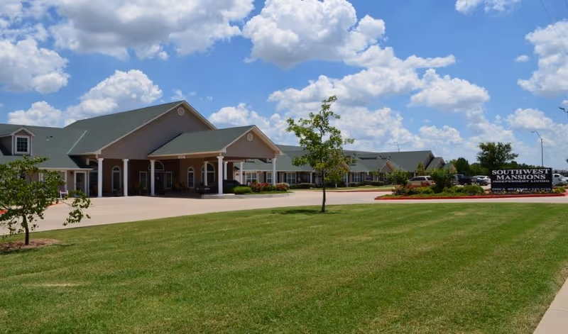Exterior view of Southwest Mansions Senior Independent Living facility showing a single-story building with a green roof, covered entrance, well-maintained lawn, small trees, and a sign with the facility name under a partly cloudy blue sky.