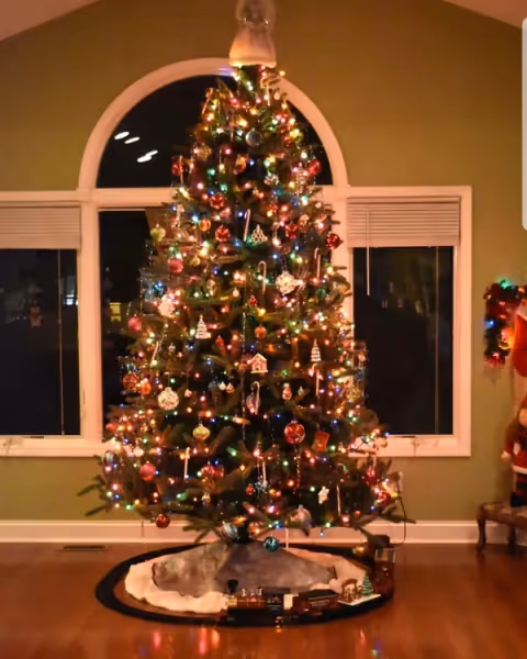 A large, decorated Christmas tree with multicolored lights stands in front of a large arched window in a living room.
