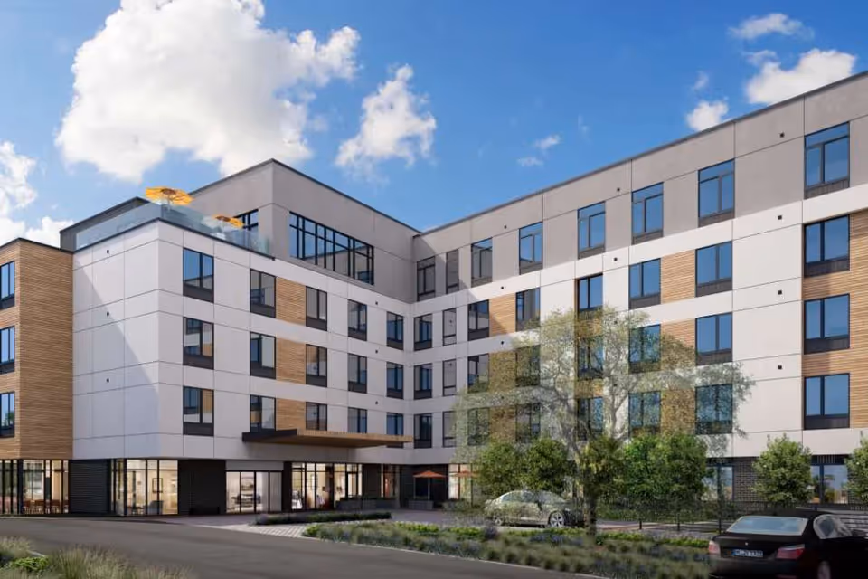 Modern five-story apartment building with large windows, an entrance canopy, landscaping, and parked cars under a blue sky.