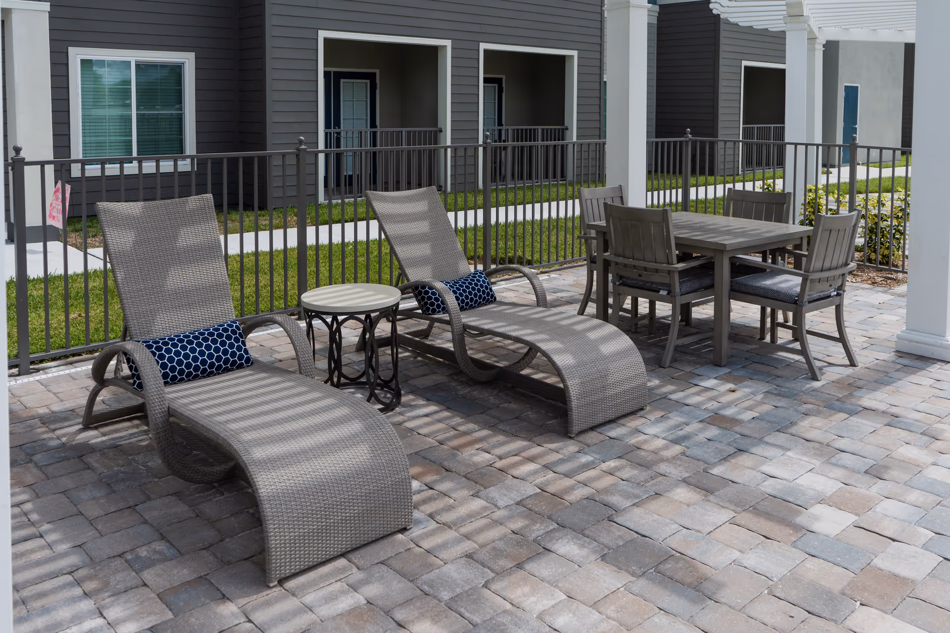 Outdoor patio area with two gray wicker chaise lounges with blue patterned pillows, a small round side table between them, and a dining table with four chairs on a paved stone floor. The area is enclosed by a black metal fence and is adjacent to a building with dark siding and white trim.