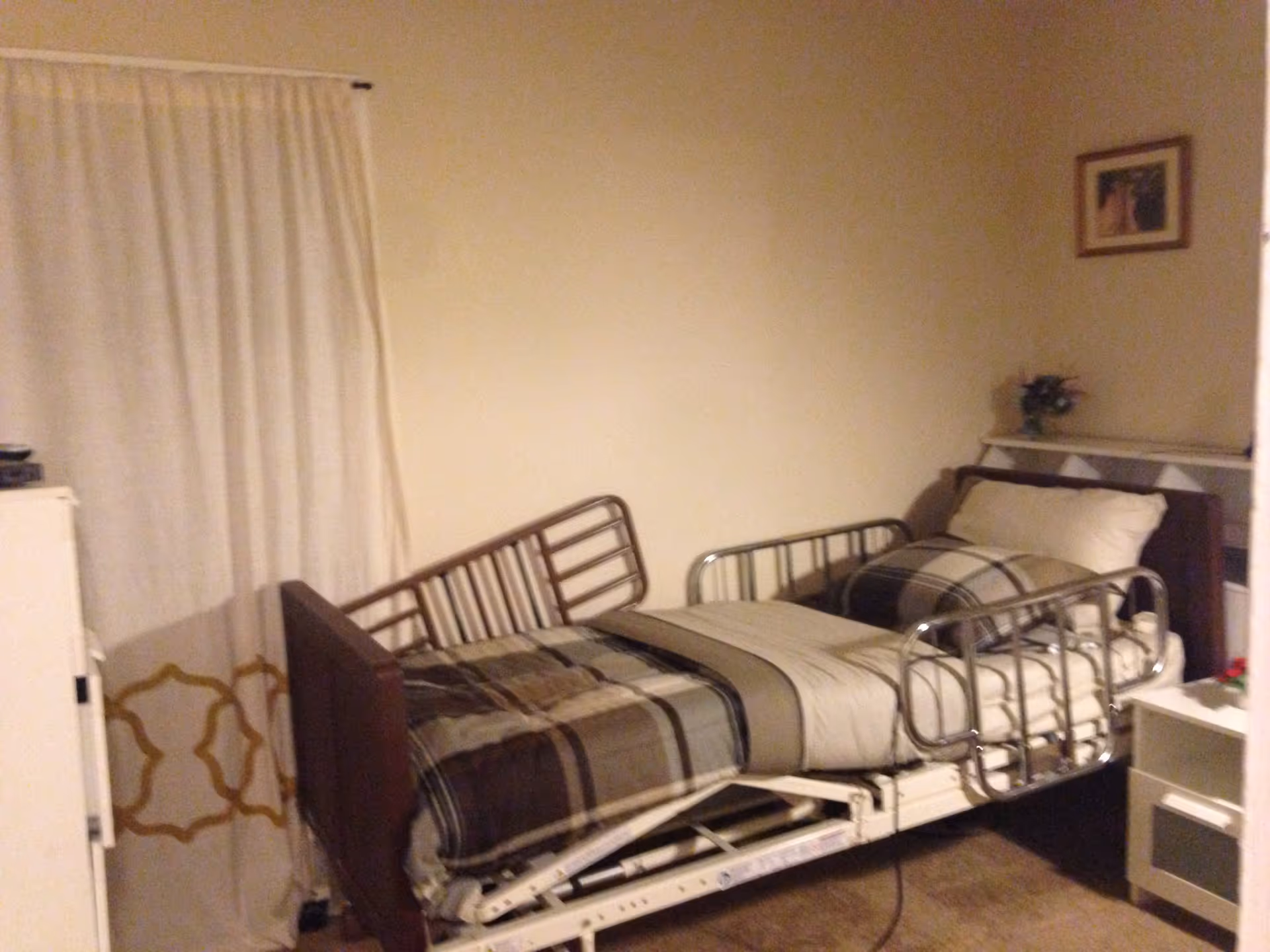 A small bedroom with a hospital-style adjustable bed made with beige and brown plaid bedding. The bed has metal side rails and is positioned against a beige wall. There is a white curtain with a gold pattern on the left side, a small white cabinet next to the bed, and a framed picture hanging on the wall above a small shelf with a flower arrangement.