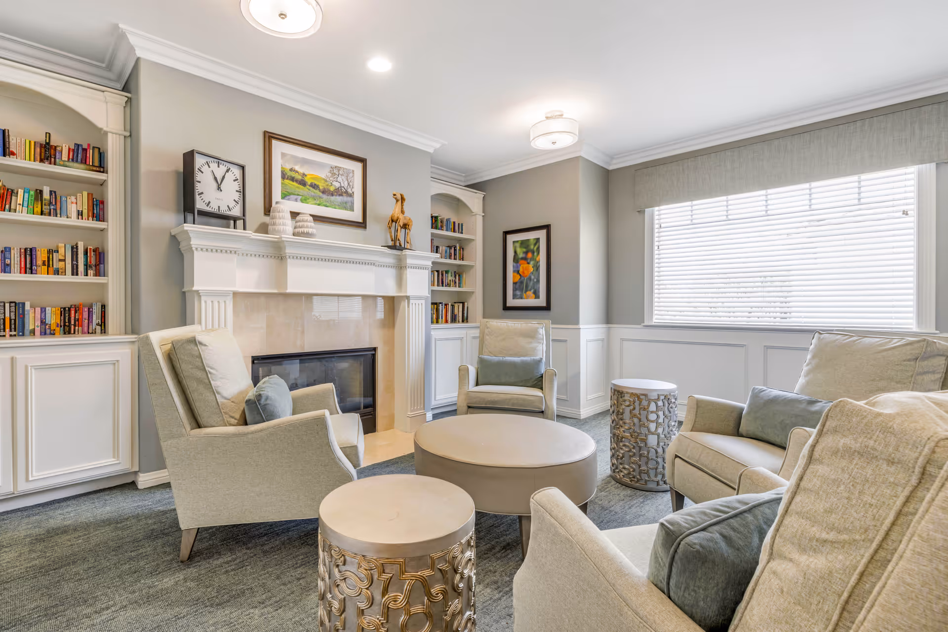 A cozy living room with four beige armchairs arranged around a round beige ottoman and two decorative side tables. The room features a white fireplace with a clock, decorative items, and a framed painting above it. Built-in bookshelves filled with books flank the fireplace. A large window with blinds allows natural light to fill the space, and the walls are painted light gray with white wainscoting.