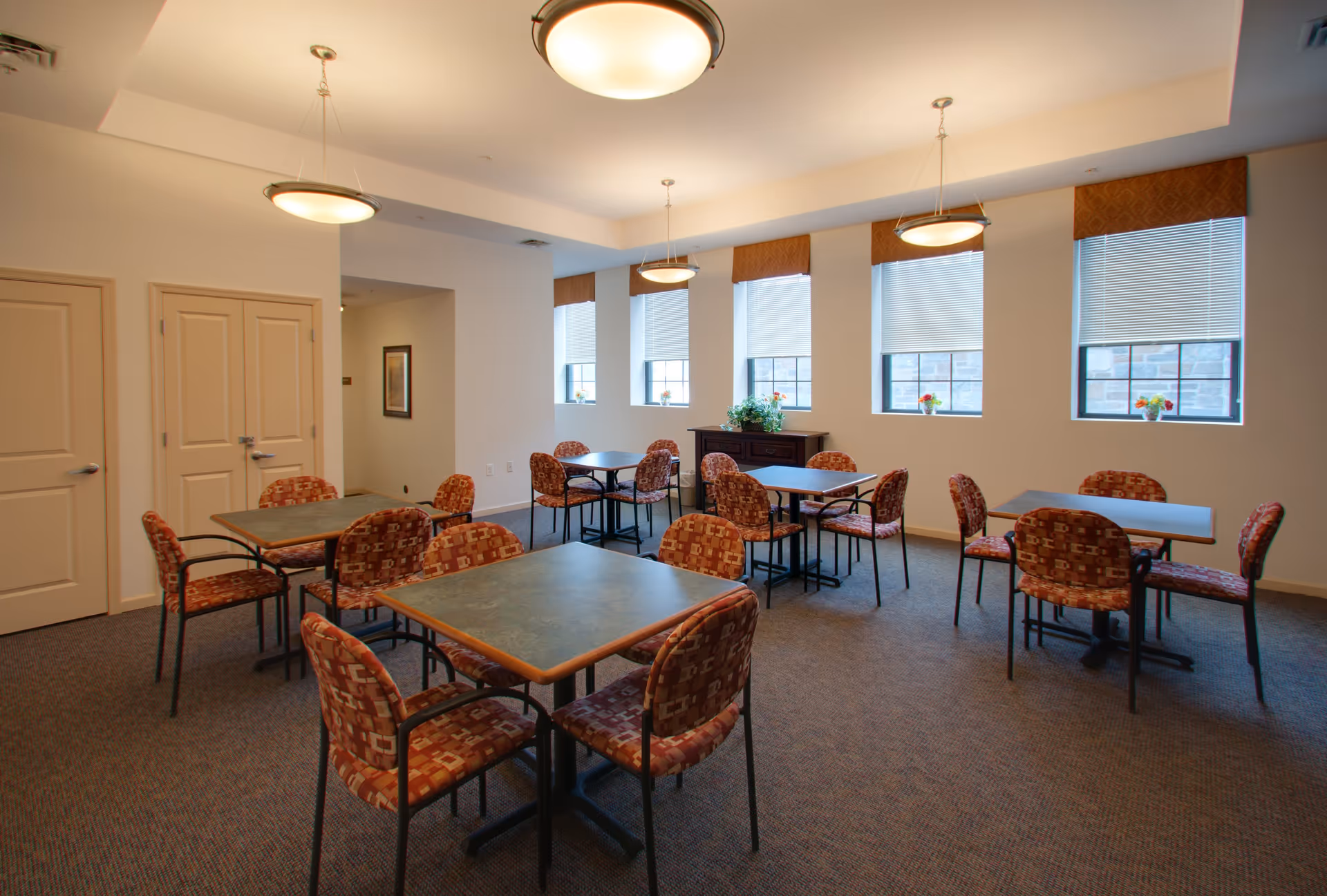 A well-lit dining room with several square tables and patterned chairs arranged neatly. The room has four windows with blinds partially drawn and small flower arrangements on the windowsills. There are three ceiling lights providing warm illumination, and a wooden cabinet with a plant on top is positioned against the far wall.